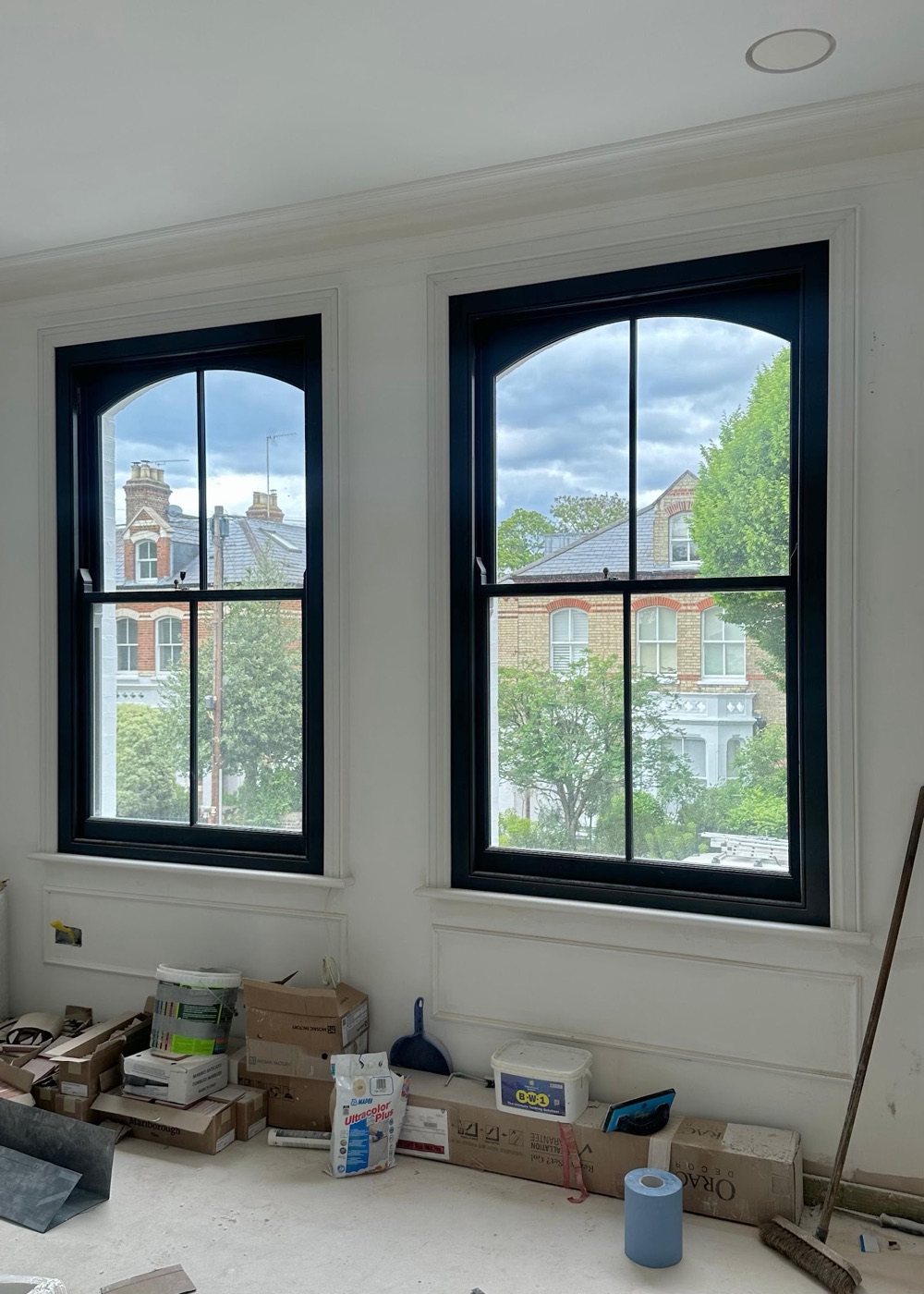 A pair of arched-head timber sash windows painted deep navy, newly fitted in a light-filled London room with rooftops beyond.