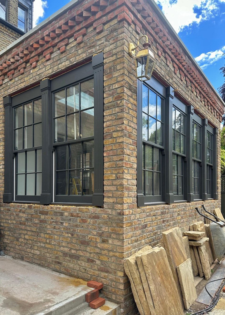 A run of tall dark-framed timber windows with slim glazing bars set into a new brick extension, in a steel-look style.